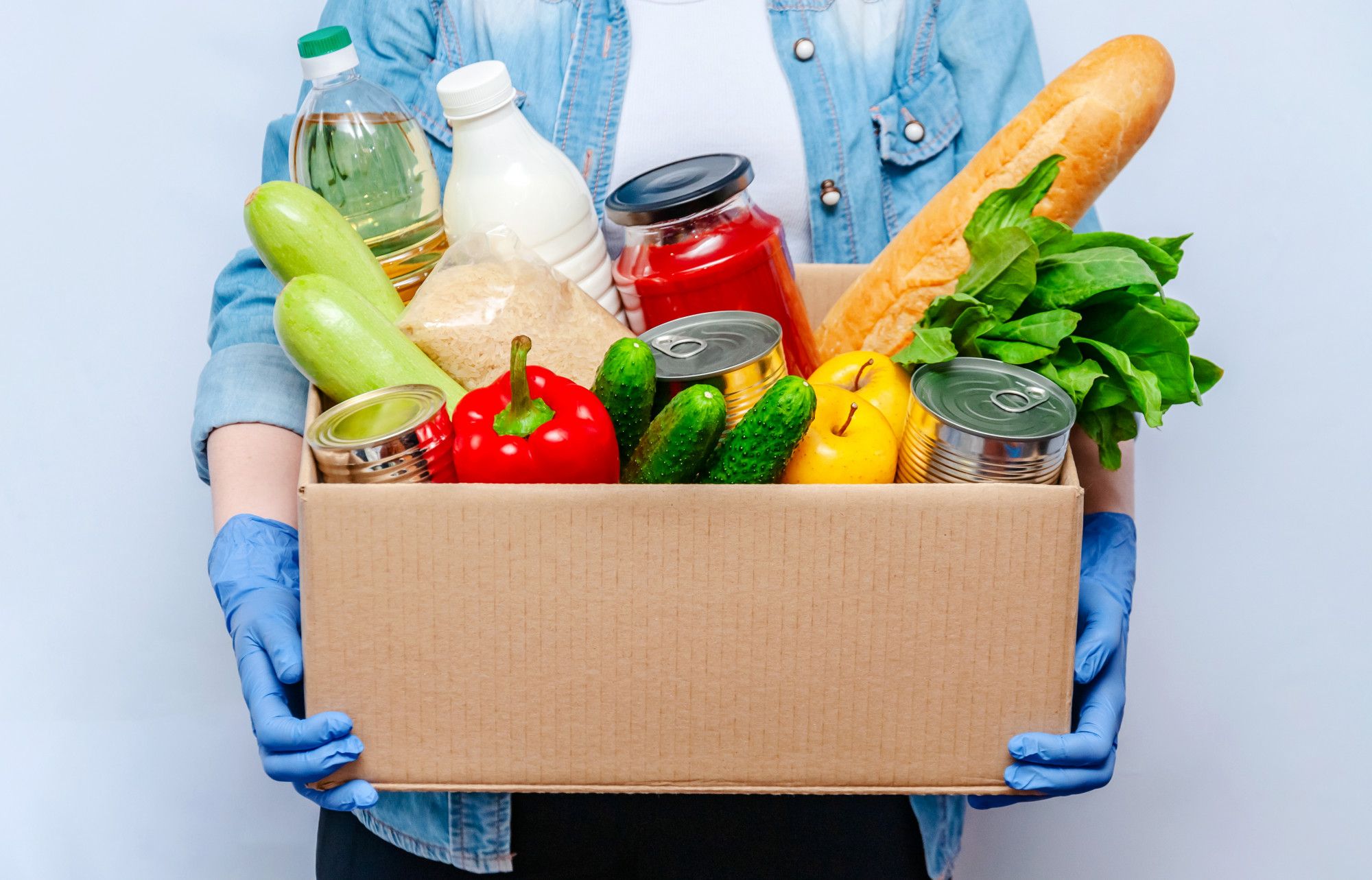woman-gloves-holding-donation-box-food-supplies-people-isolation-essential-goods-oil-canned-food-cereals-milk-vegetables-fruit